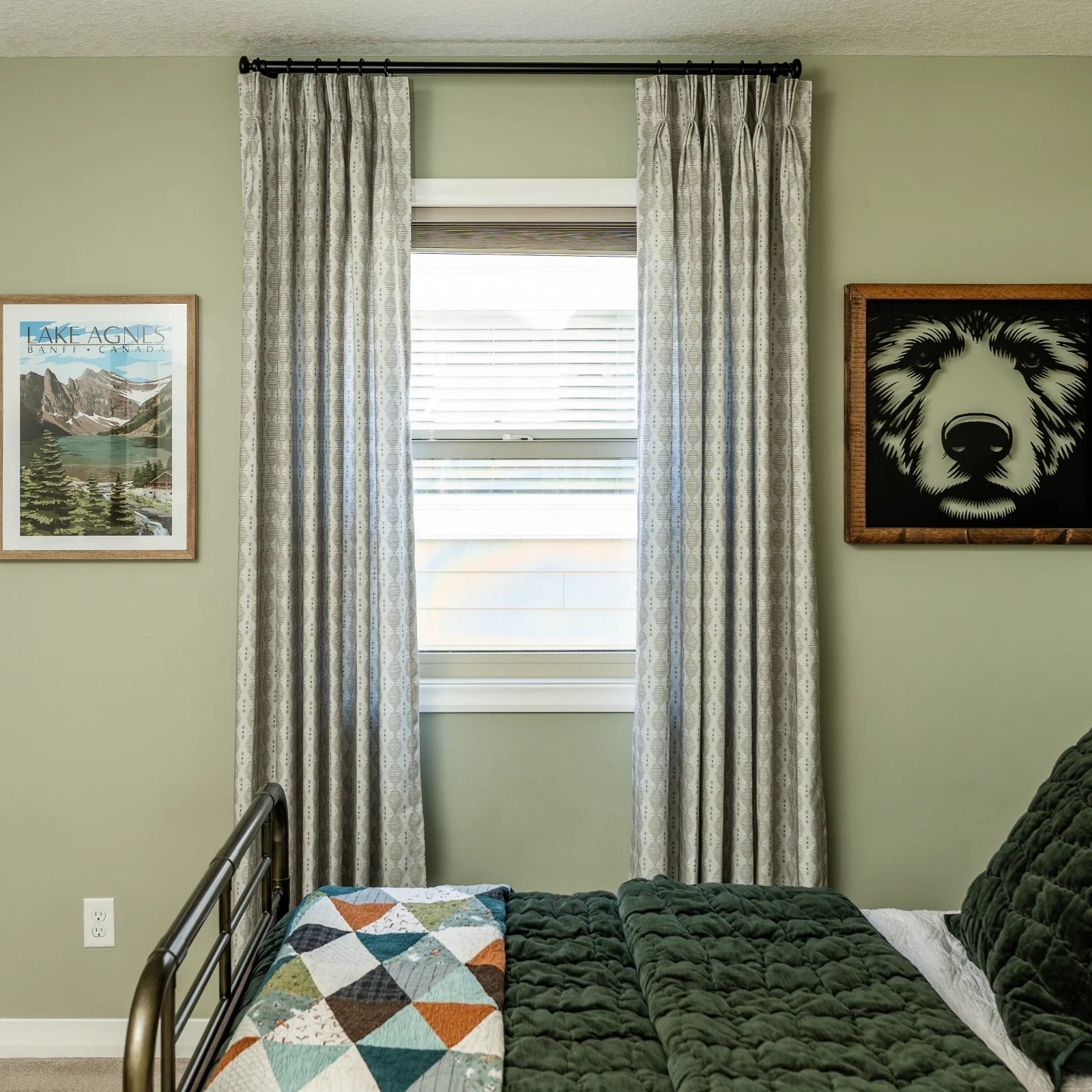 Photograph of a cozy bedroom featuring sage green walls a bed with a dark green quilted duvet and a colorful geometric quilt patterned grey curtains flanking a window a Lake Agnes Banff Canada poster and a framed bear face art print