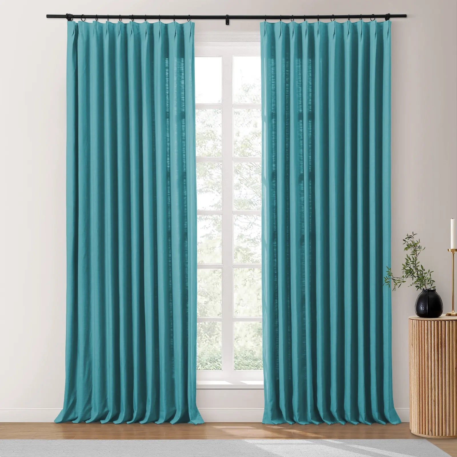 A pair of pleated teal curtains on a black rod frame a bright window in a room featuring a wooden fluted side table with a black vase and greenery