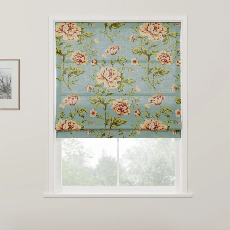 Fabric roman blind featuring a vintage floral pattern with large peach and yellow roses and green leaves on a teal blue background, partially covering a white window with a view of trees