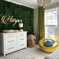 Photograph of a modern child's nursery featuring a large artificial greenery wall with a gold 'Harper' sign, a white changing dresser, a woven elephant basket, and a bright yellow swivel chair with a blue textured pillow