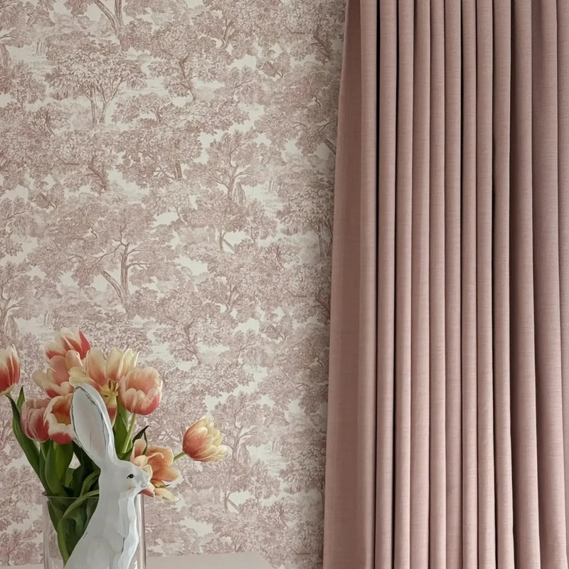 Photograph of an elegant interior featuring a clear vase with a white bunny figurine and vibrant pink and peach tulips against a light brown toile tree pattern wallpaper next to a dusty rose pleated curtain
