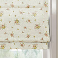 Creamy white textured Roman blind featuring a vintage-style pattern of scattered pink and yellow floral clusters and green leaves, partially raised in a white window frame to reveal blurry green trees outside