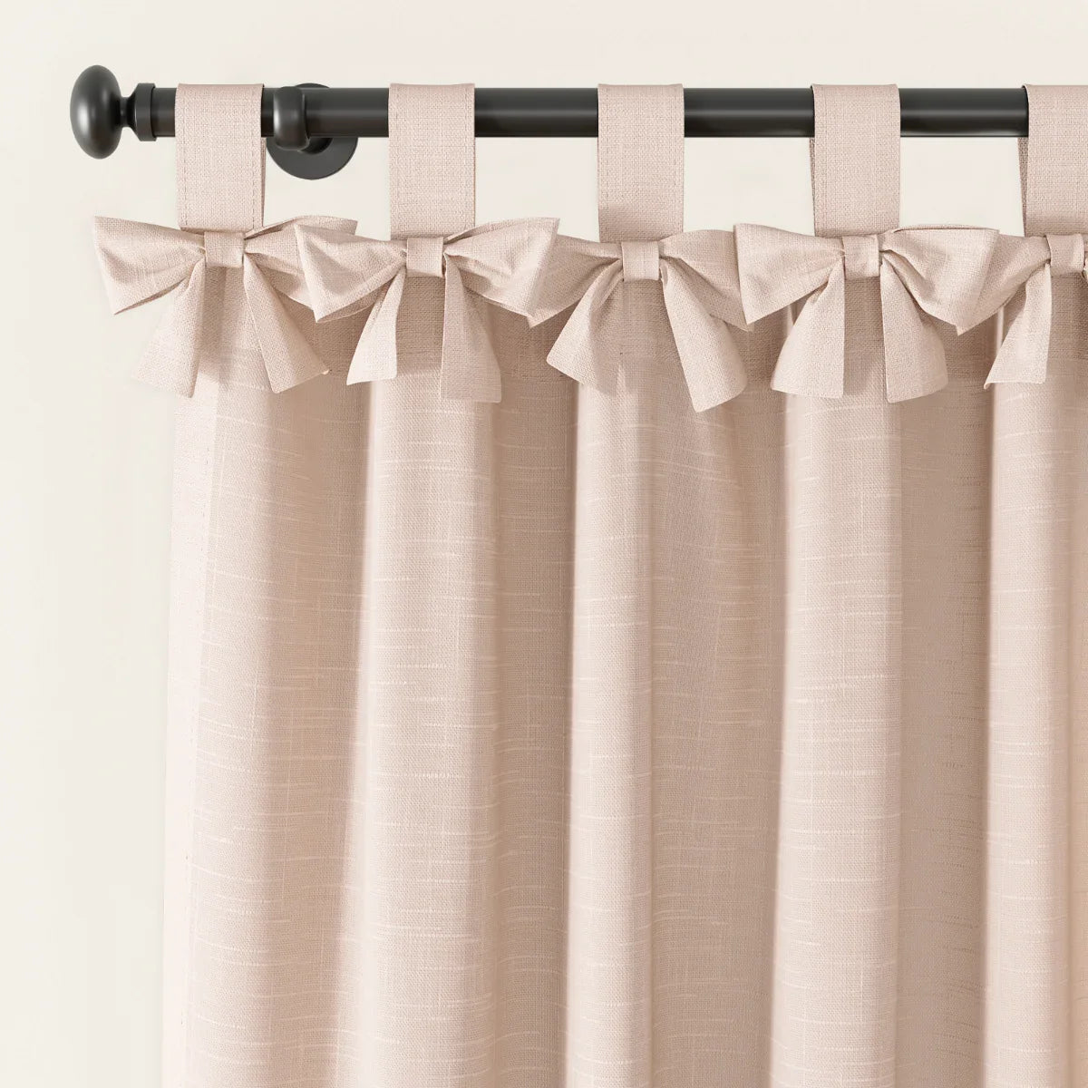 A blush pink textured curtain with decorative bow tie top tabs hanging on a black rod with a round finial