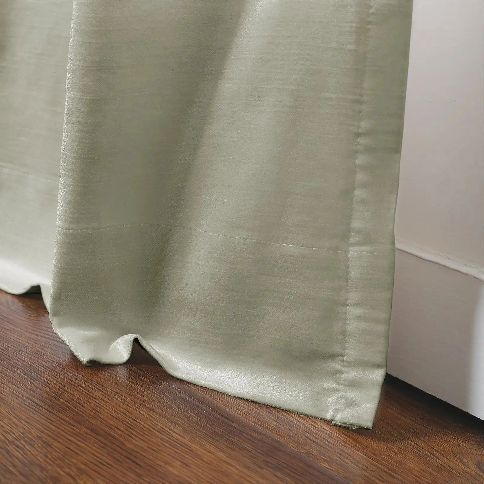 Light sage green fabric curtain with subtle texture and visible vertical seams pooling gently on a warm brown hardwood floor