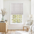 A bright minimalist bedroom interior featuring a window with a light grey striped Roman shade, a natural wood dresser topped with a pale green vase and dried floral branches, and a cream upholstered chair with a soft white pillow and throw