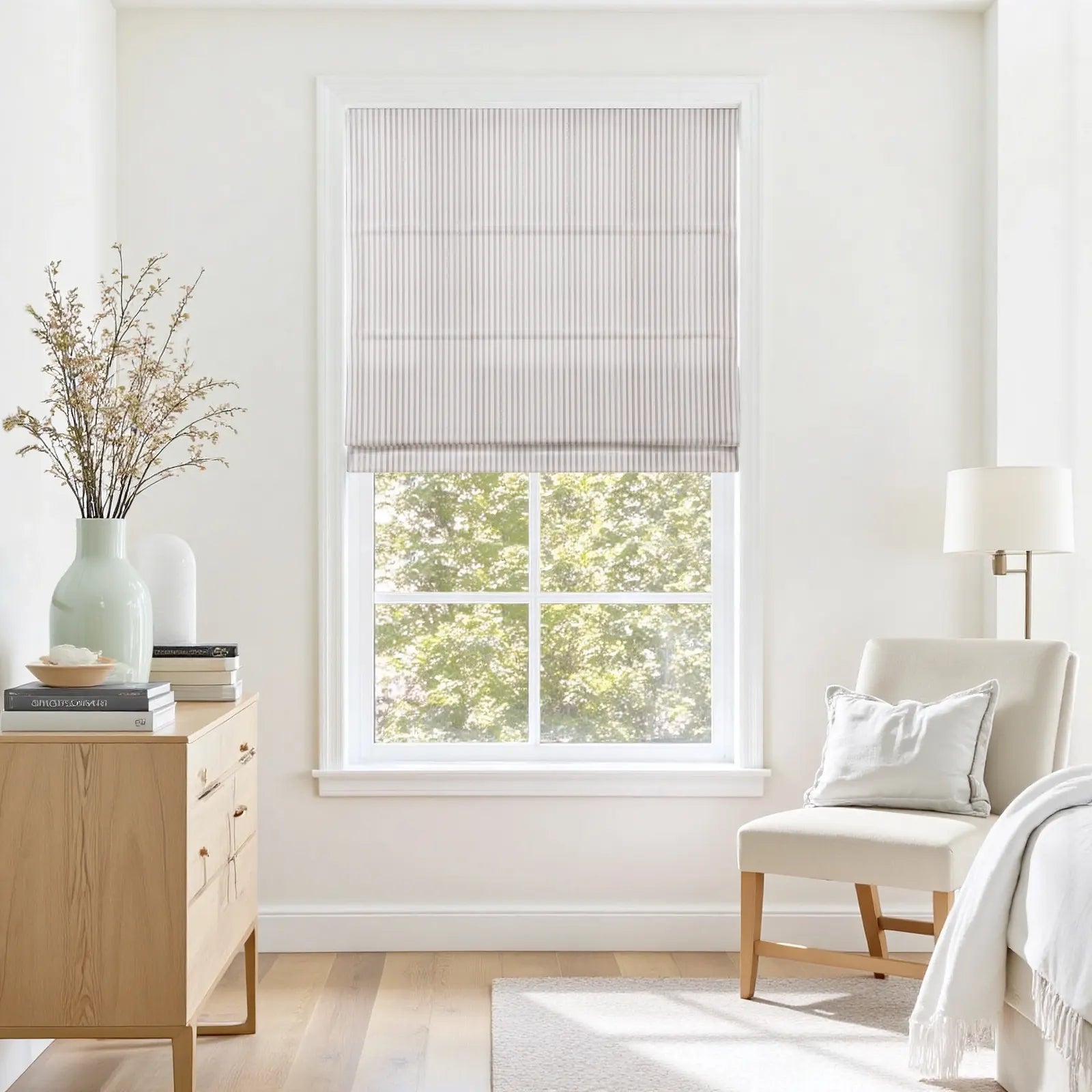 A bright minimalist bedroom interior featuring a window with a light grey striped Roman shade, a natural wood dresser topped with a pale green vase and dried floral branches, and a cream upholstered chair with a soft white pillow and throw