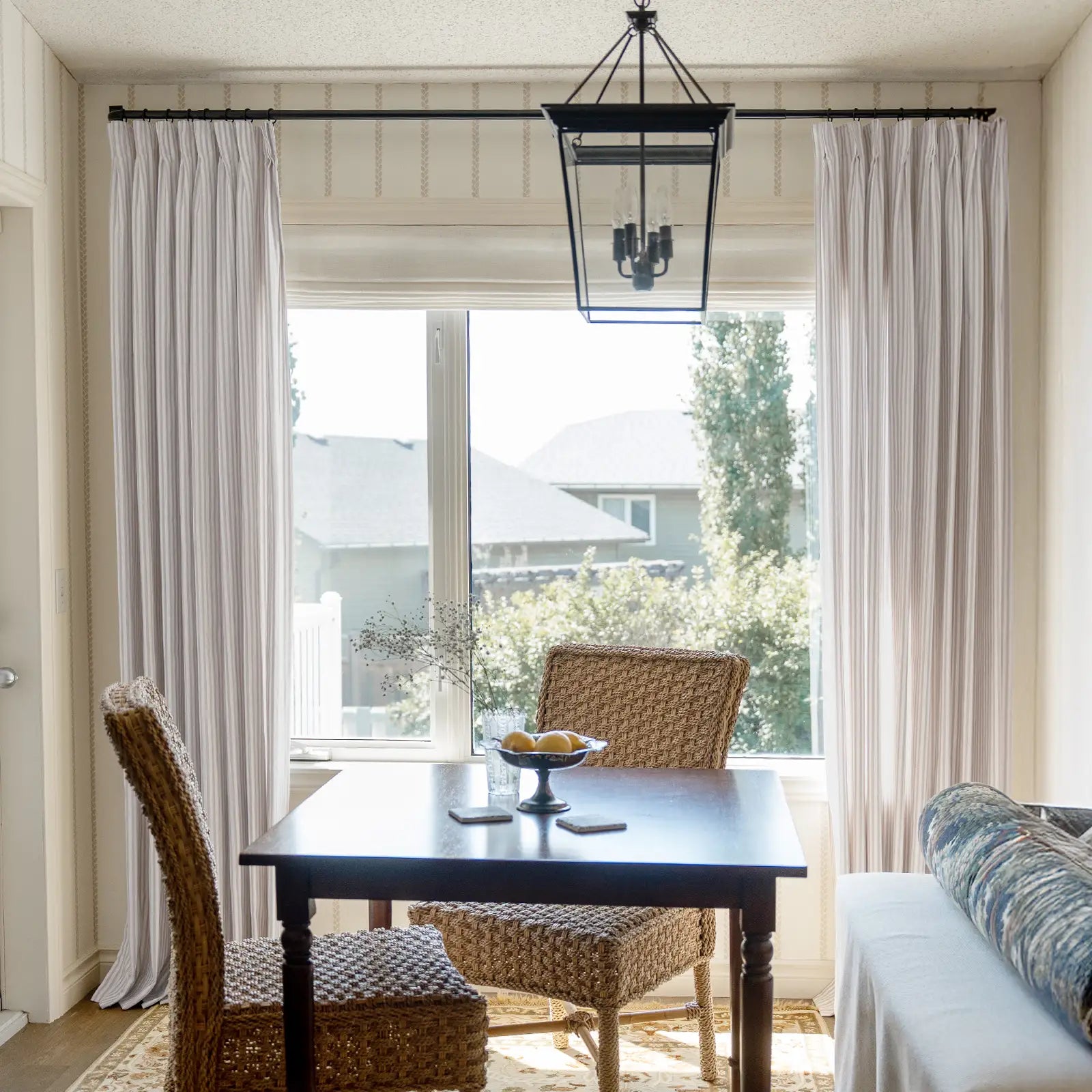 Interior photograph of a bright dining nook with a dark wood table, woven wicker chairs, a black lantern pendant light, and striped curtains framing a sunny window with a bowl of lemons