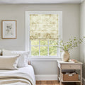 A serene bedroom interior featuring a window with a cream and green floral Roman shade, white linen bedding with a woven accent pillow, and a natural wood nightstand holding a cream vase with fresh green branches beside a framed botanical print