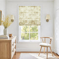 Photograph of a bright and airy room interior featuring a window with a delicate floral Roman shade revealing lush green trees a light wood dresser holding a tall white vase of vibrant yellow flowers and stacked books alongside a simple wooden chair and elegant wall sconces