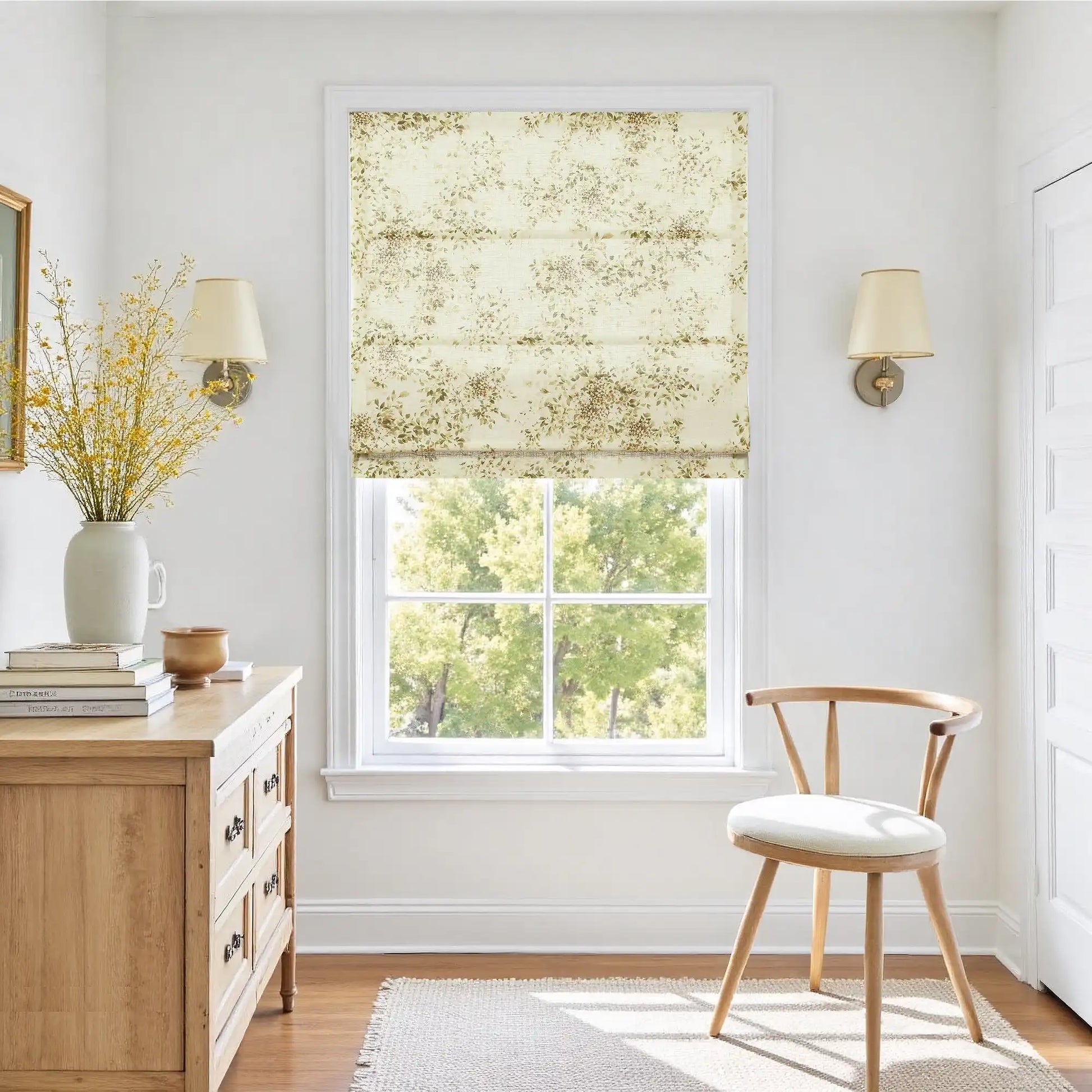 Photograph of a bright and airy room interior featuring a window with a delicate floral Roman shade revealing lush green trees a light wood dresser holding a tall white vase of vibrant yellow flowers and stacked books alongside a simple wooden chair and elegant wall sconces