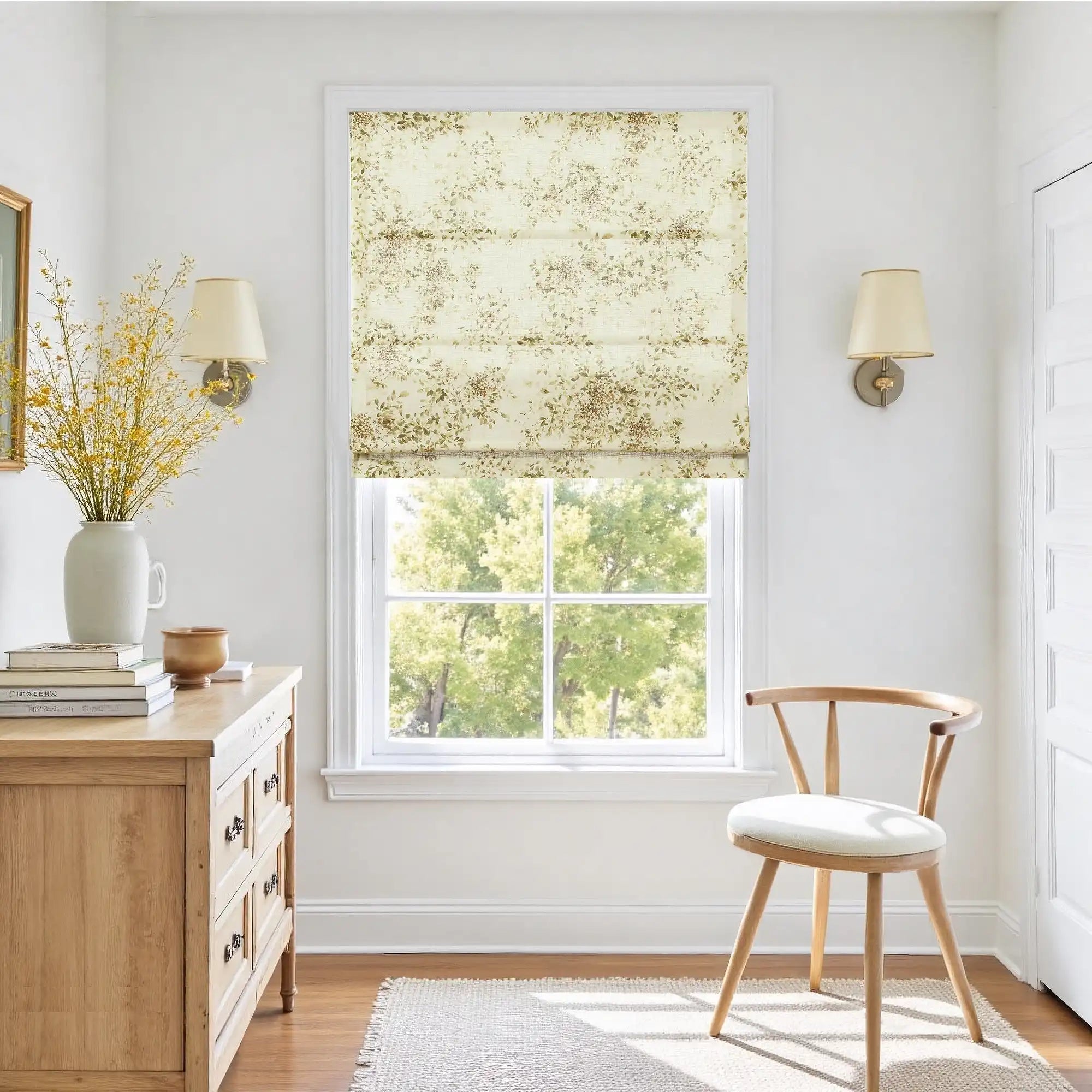 Photograph of a bright and airy room interior featuring a window with a delicate floral Roman shade revealing lush green trees a light wood dresser holding a tall white vase of vibrant yellow flowers and stacked books alongside a simple wooden chair and elegant wall sconces