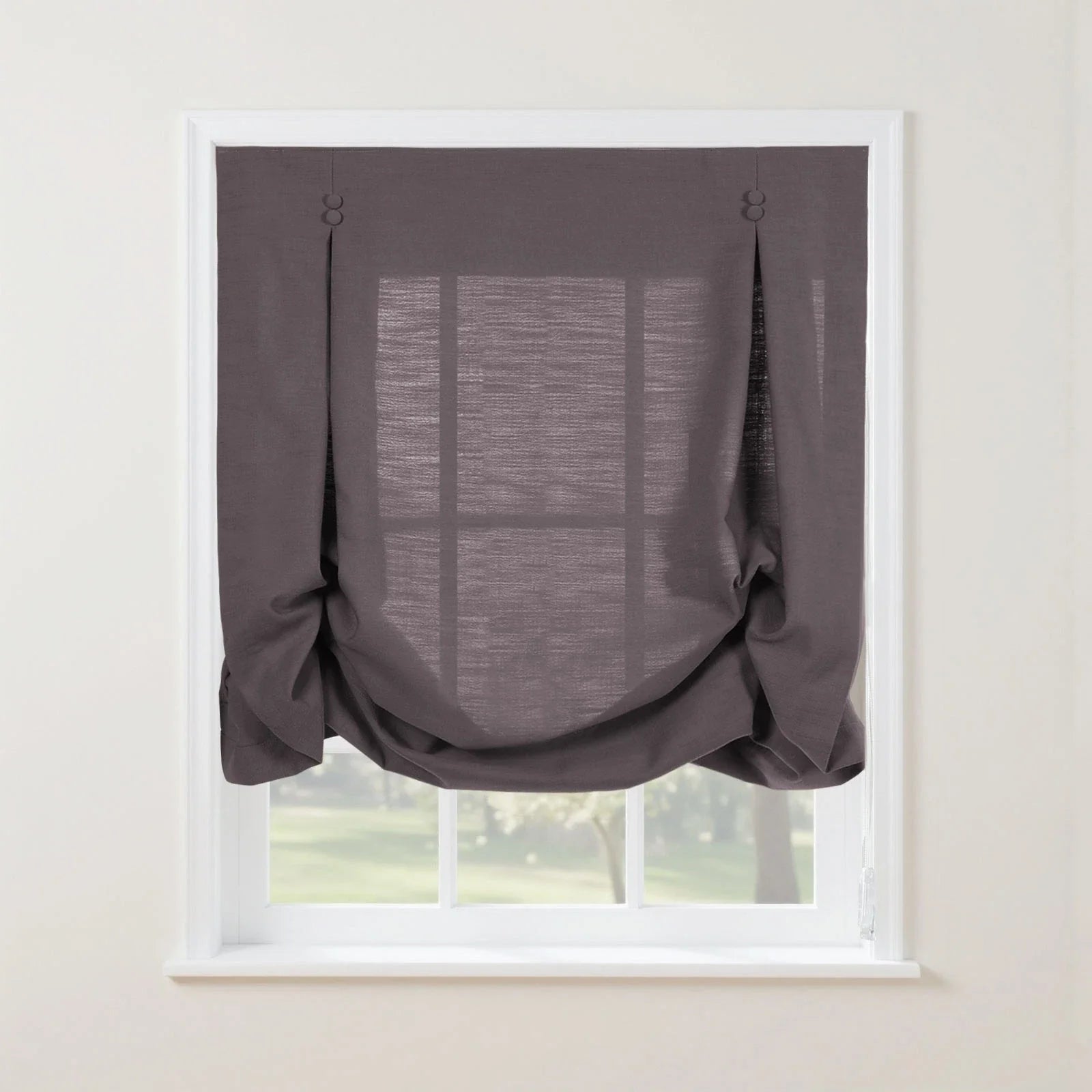 Dark gray linen balloon shade with decorative fabric button loops and a softly gathered bottom covering a white window frame revealing blurred green trees