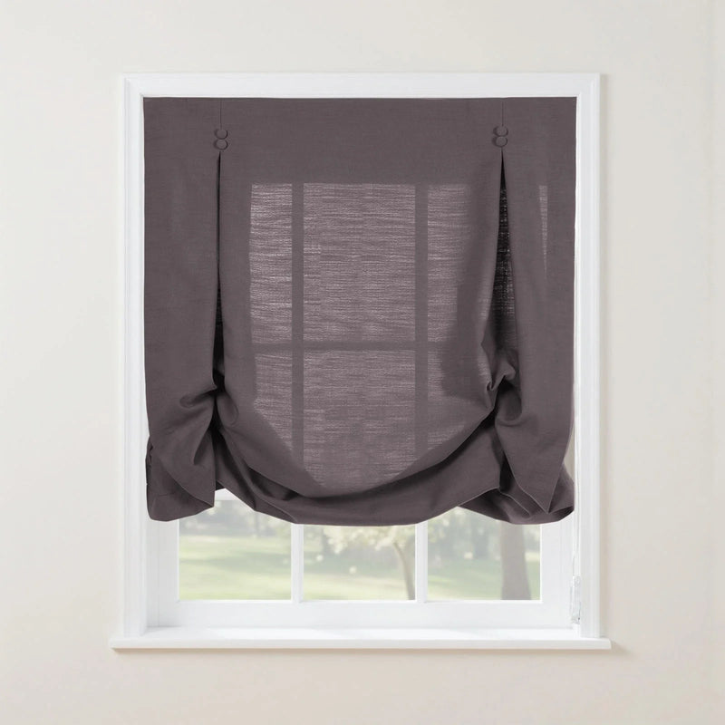 Dark gray linen balloon shade with decorative fabric button loops and a softly gathered bottom covering a white window frame revealing blurred green trees