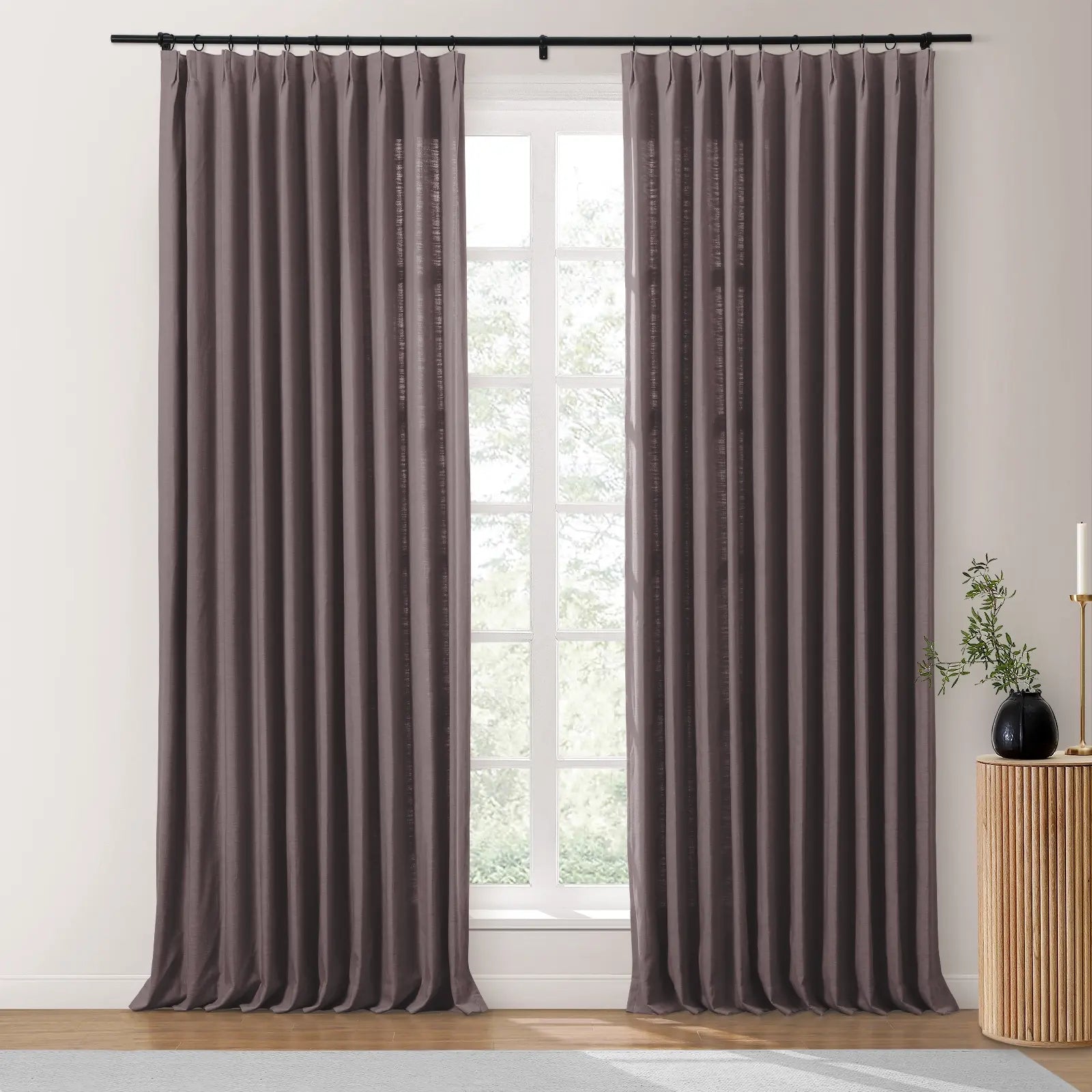 Photograph of a window in a room adorned with long taupe pinch-pleat curtains on a black rod, revealing lush green trees outside, complemented by a fluted wooden side table holding a black vase with foliage and a brass candle holder
