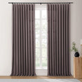 Photograph of a window in a room adorned with long taupe pinch-pleat curtains on a black rod, revealing lush green trees outside, complemented by a fluted wooden side table holding a black vase with foliage and a brass candle holder