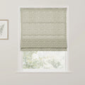 Photograph of an interior window featuring a beige roman blind with a dense sage green floral pattern, a white window frame, and a gold-framed botanical print of a single green leaf on the left