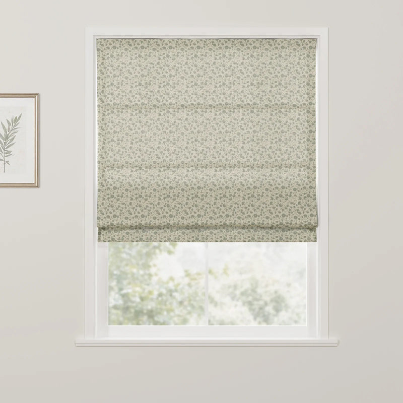 Photograph of an interior window featuring a beige roman blind with a dense sage green floral pattern, a white window frame, and a gold-framed botanical print of a single green leaf on the left