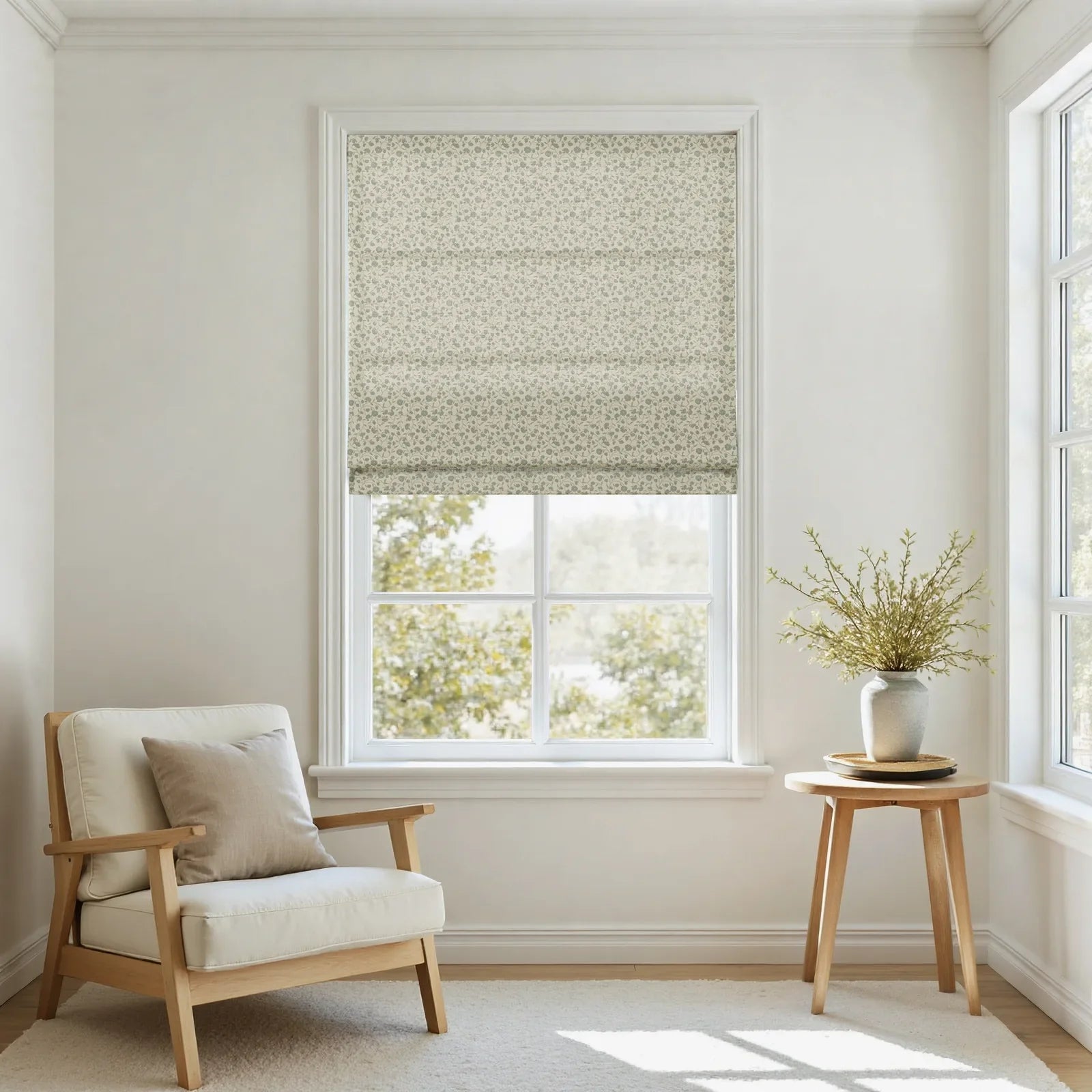 Photograph of a bright room interior with a partially lowered green floral Roman blind, a cream upholstered armchair with a beige cushion, and a wooden side table holding a gray vase filled with green foliage