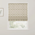 Photograph of an interior window featuring a cream Roman blind with a subtle repeating floral pattern in muted green partially revealing a blurred green outdoor scene with a gold-framed botanical print on the left white wall