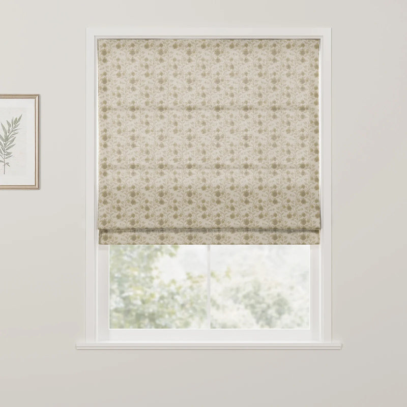Photograph of an interior window featuring a cream Roman blind with a subtle repeating floral pattern in muted green partially revealing a blurred green outdoor scene with a gold-framed botanical print on the left white wall