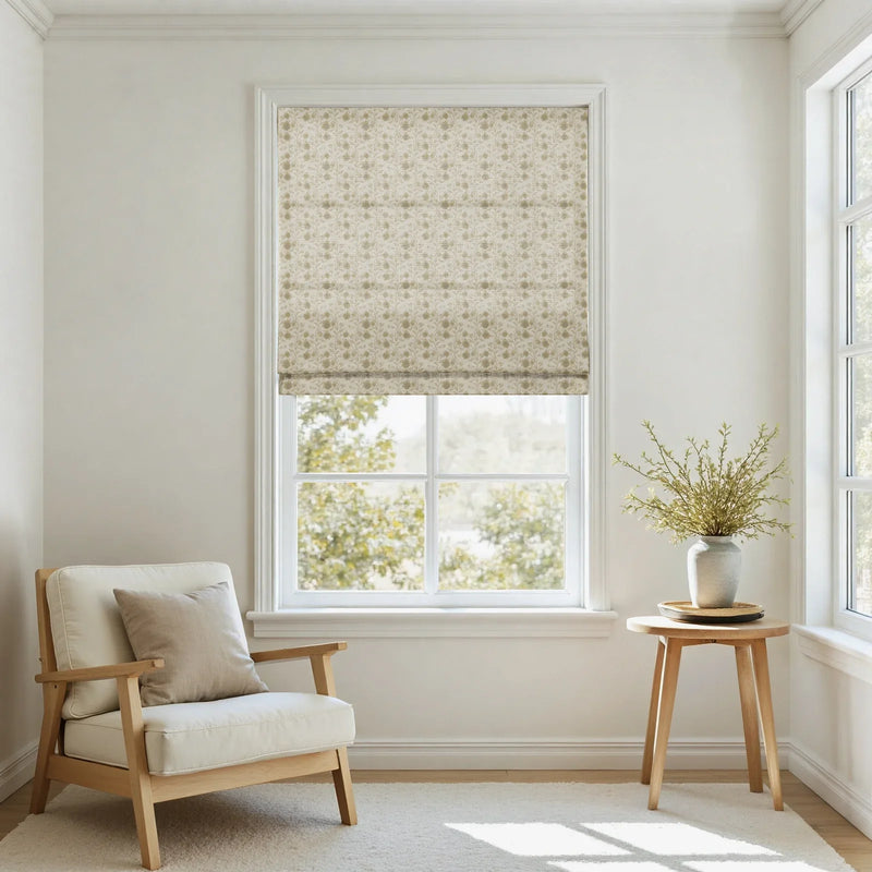 An interior room with a window featuring a beige floral patterned Roman shade, a light wooden armchair with cream cushions, and a wooden side table displaying a gray vase with green foliage