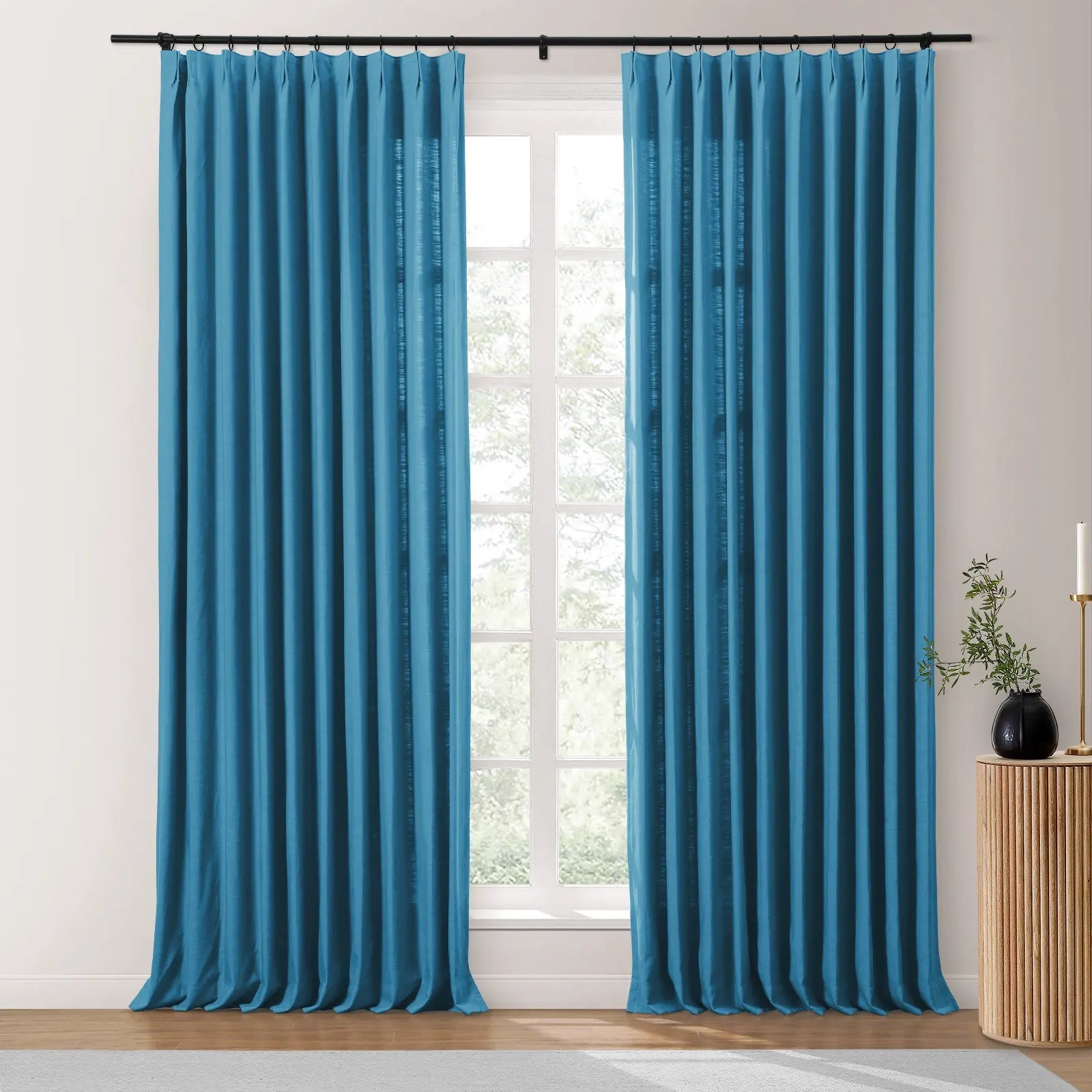 Vibrant teal blue pinch pleat curtains hang from a black rod flanking a window with an outdoor tree view, beside a fluted wood pedestal displaying a black vase with greenery and a brass candlestick