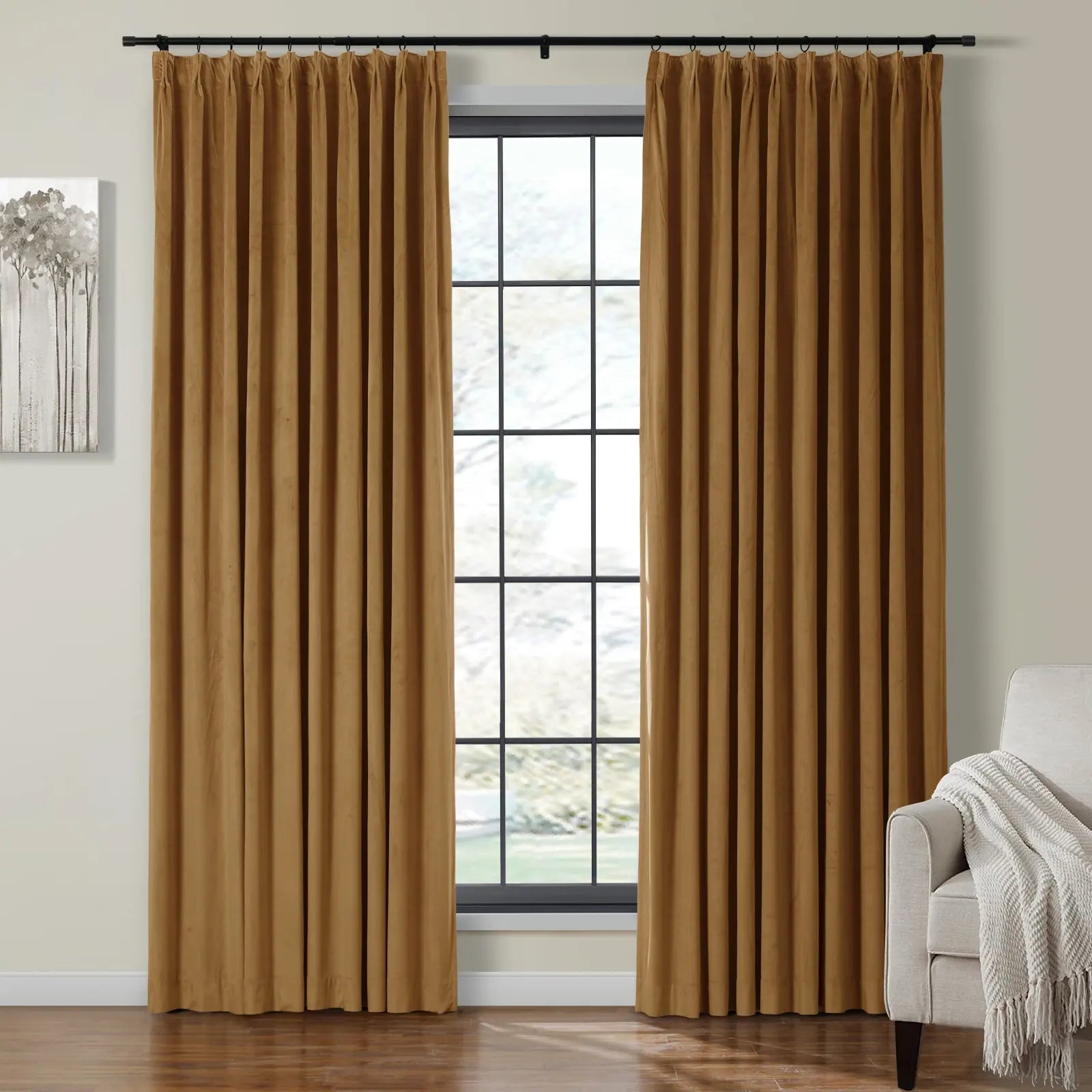 A pair of warm caramel pleated curtains framing a large grid-style window in a cozy room with abstract tree art and a light gray armchair draped with a white throw