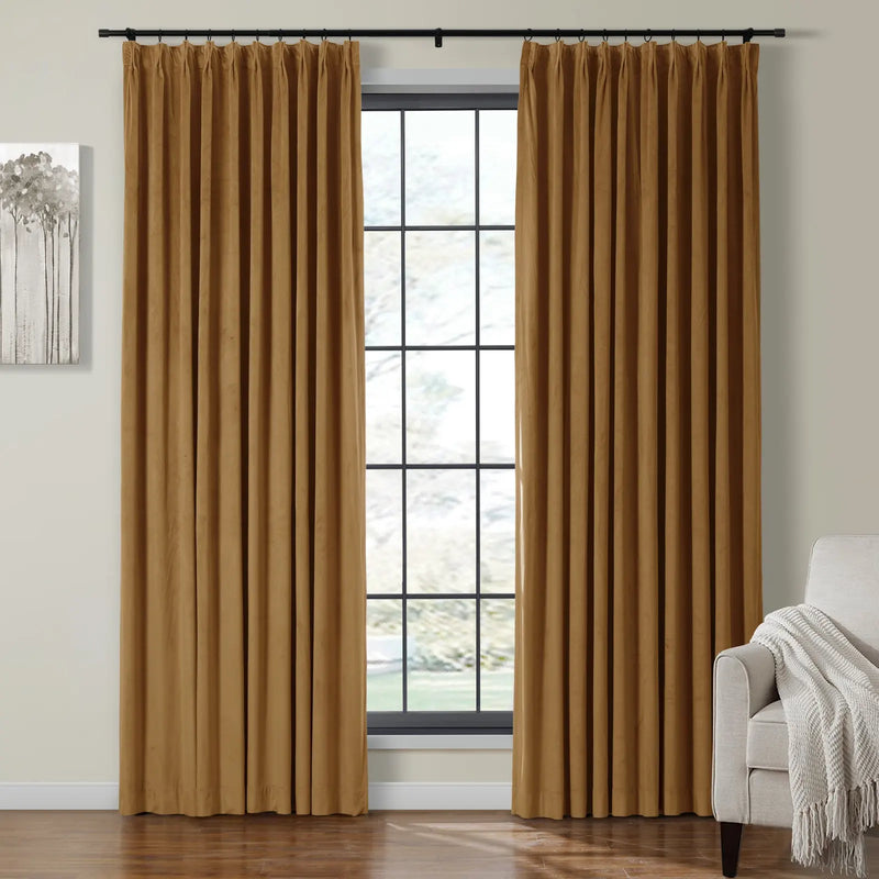 A pair of warm caramel pleated curtains framing a large grid-style window in a cozy room with abstract tree art and a light gray armchair draped with a white throw