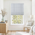 A bright bedroom interior featuring a window with a blue and white striped Roman shade, a light wood dresser with a mint green vase of dried branches, and a cream accent chair with a white throw
