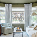 A warm living room with olive green walls, a bay window flanked by light gray striped drapes over roman shades, a cozy striped armchair, a white sofa, and a coffee table with a glowing candle