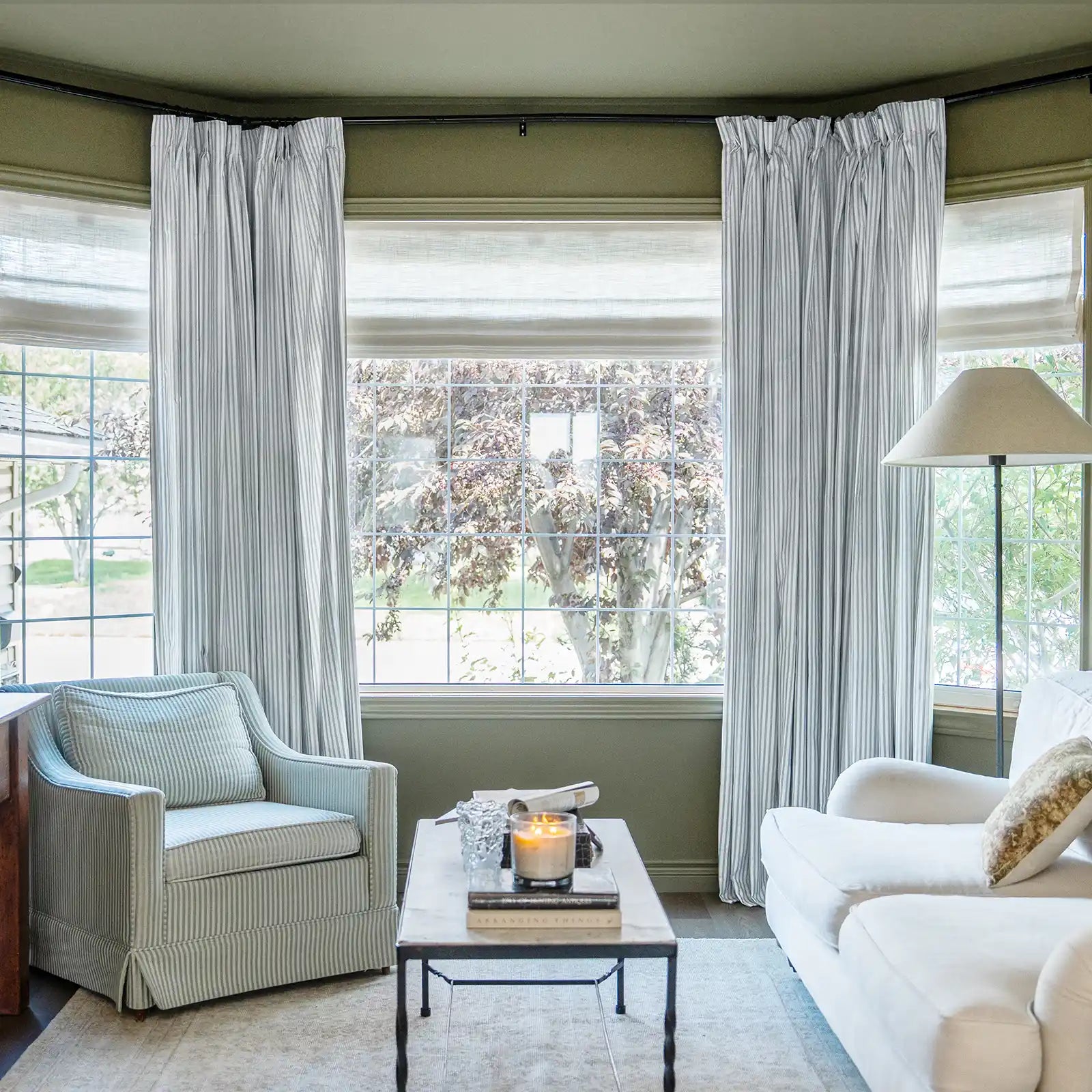 A warm living room with olive green walls, a bay window flanked by light gray striped drapes over roman shades, a cozy striped armchair, a white sofa, and a coffee table with a glowing candle