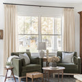Sand Beige Liz Linen Pleated Curtains framing a green sofa beneath a wide sunlit window