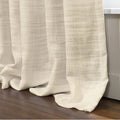 Cream linen curtains with a visible textured weave forming soft folds that gracefully pool onto a dark wood floor beside a white baseboard