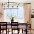 Photograph of an elegant dining room interior with a large window overlooking a bare tree landscape, featuring a black geometric chandelier, a navy blue and white floral tablecloth, and a small dog resting under the table