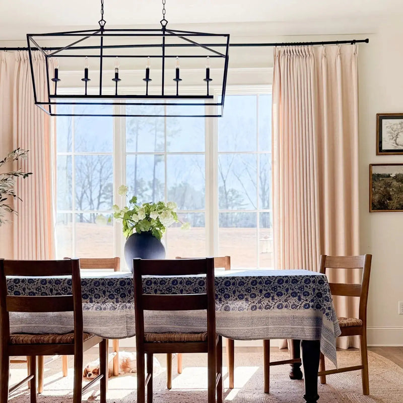 Photograph of an elegant dining room interior with a large window overlooking a bare tree landscape, featuring a black geometric chandelier, a navy blue and white floral tablecloth, and a small dog resting under the table