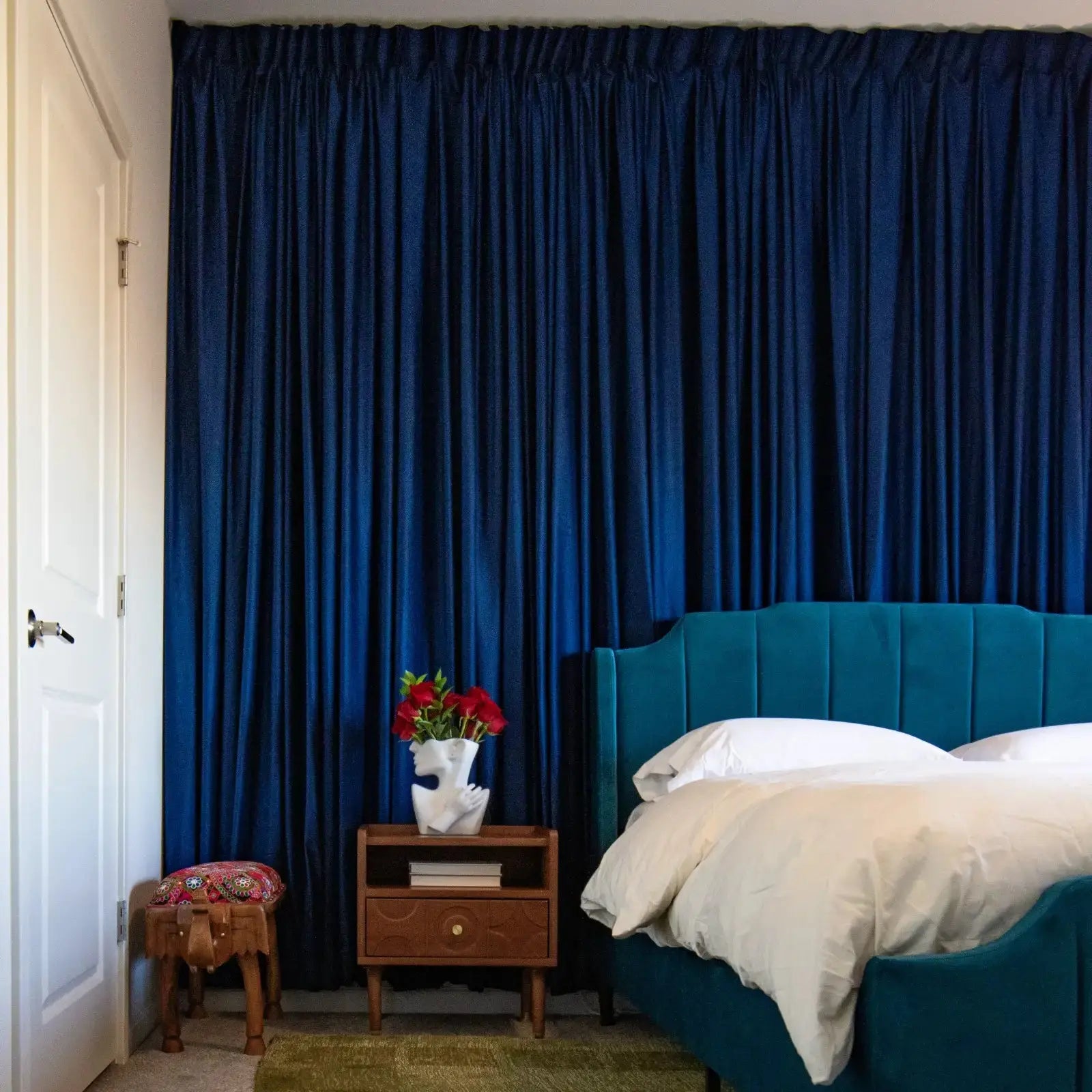 A vibrant bedroom interior showcasing floor-to-ceiling dark blue velvet curtains, a teal upholstered bed with white bedding, and a unique white head-shaped vase filled with red roses on a wooden nightstand