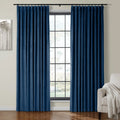 A pair of deep royal blue velvet pleated curtains hanging on a black rod over a large window with black grid panes in a modern living room featuring a minimalist tree painting and a cozy beige armchair draped with a white throw blanket