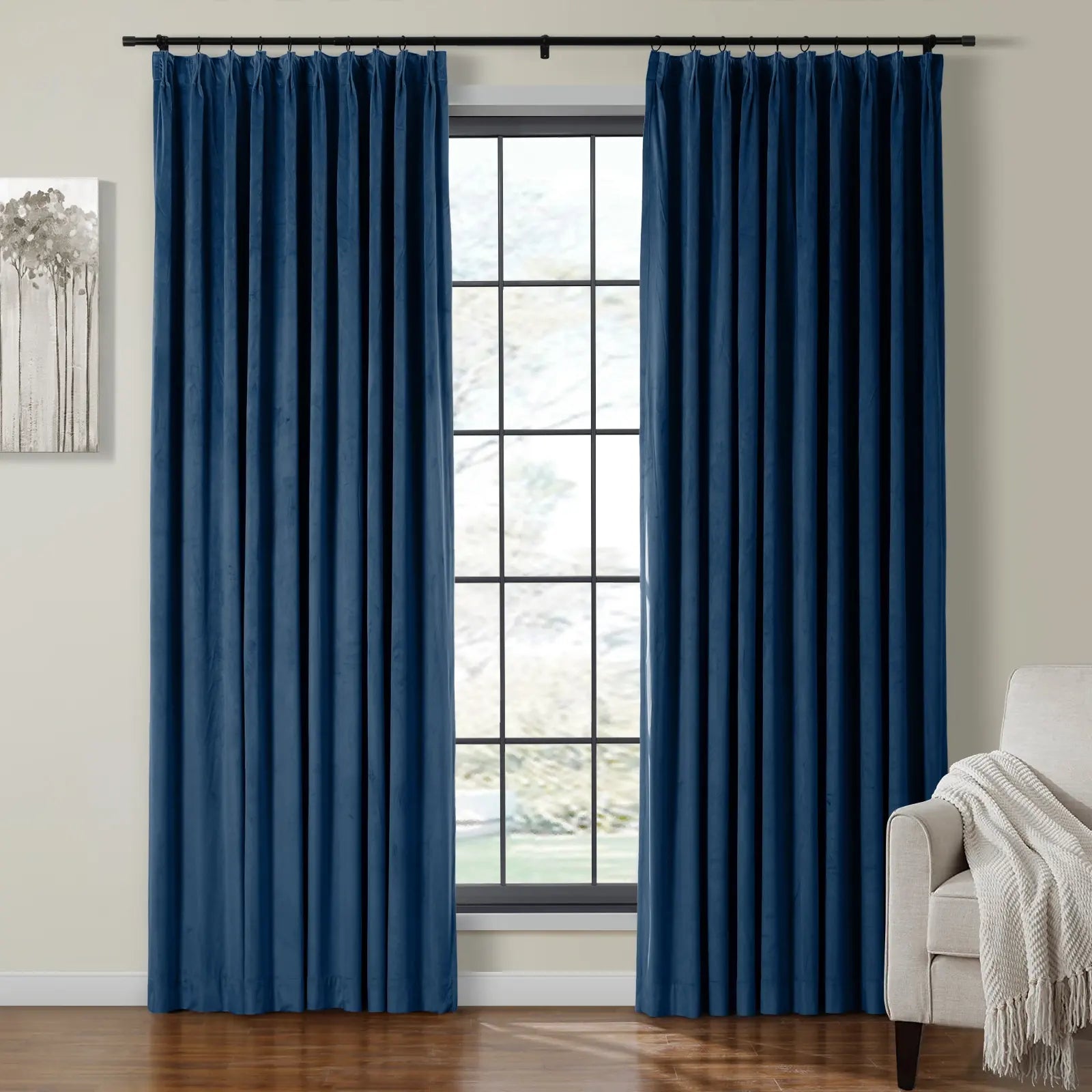 A pair of deep royal blue velvet pleated curtains hanging on a black rod over a large window with black grid panes in a modern living room featuring a minimalist tree painting and a cozy beige armchair draped with a white throw blanket