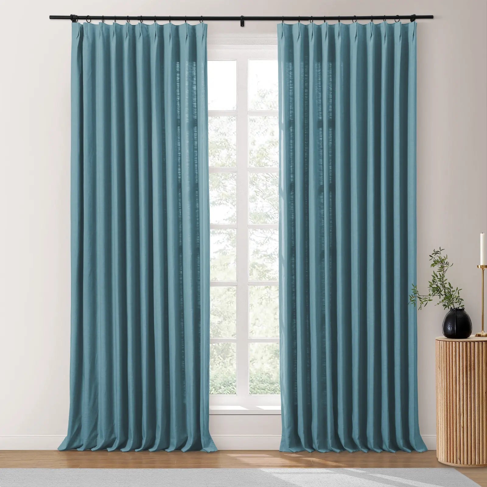 Teal pleated curtains with a black rod framing a large window with a view of bare trees, a fluted wooden side table with a black vase holding green foliage, and a gold candlestick
