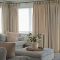 An interior living room scene featuring floor-to-ceiling beige drapes, a light blue sectional sofa with a blush pink throw pillow, and a white ottoman serving as a coffee table with a woven tray holding white flowers