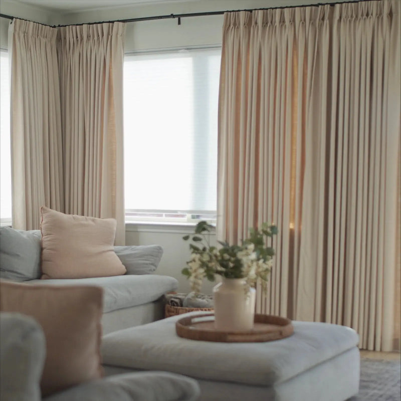 An interior living room scene featuring floor-to-ceiling beige drapes, a light blue sectional sofa with a blush pink throw pillow, and a white ottoman serving as a coffee table with a woven tray holding white flowers