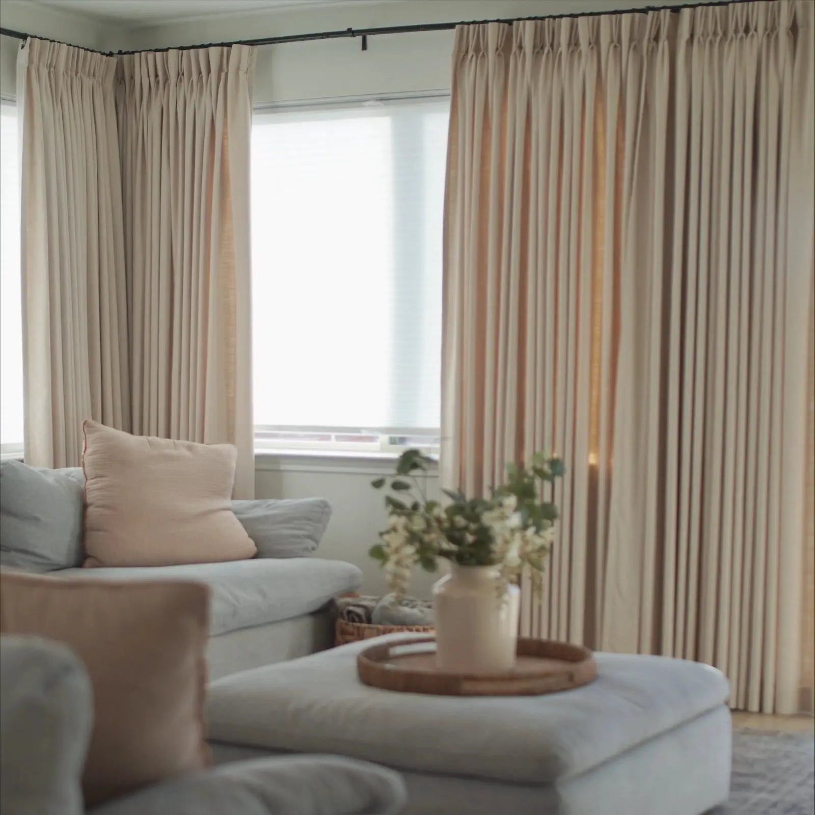 An interior living room scene featuring floor-to-ceiling beige drapes, a light blue sectional sofa with a blush pink throw pillow, and a white ottoman serving as a coffee table with a woven tray holding white flowers