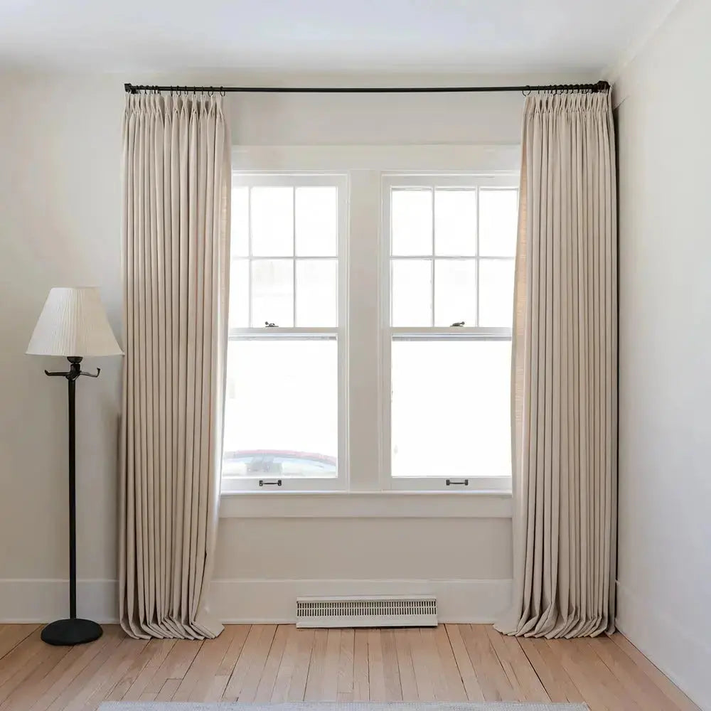 Interior shot of a bright room featuring two white multi-pane windows flanked by tall beige pleated curtains on a black rod and a black floor lamp with a pleated white blind on light hardwood floors