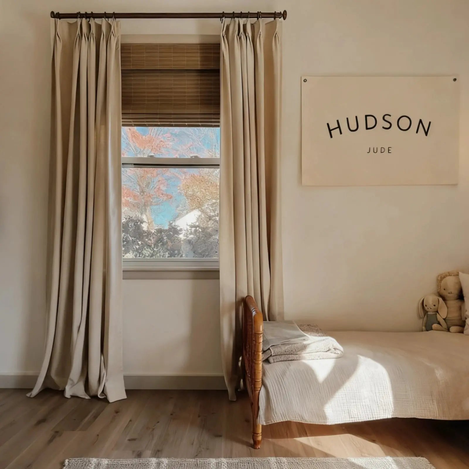 A modern child's bedroom with beige pleated curtains and bamboo blinds over a window revealing autumn trees, featuring a light wood bed with textured white bedding, two plush bunny toys, and a white canvas wall hanging boldly inscribed HUDSON JUDE