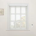 A home interior showing a window fitted with white horizontal zebra Blinds, a framed botanical print on the left wall, and a white remote control mounted on the right wall