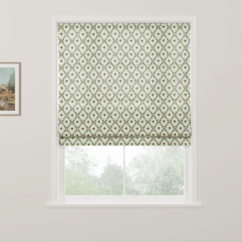 Photograph of an interior window featuring a light green Roman blind with a lattice pattern and small red floral motifs, a framed landscape painting hangs on the left wall