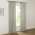 White pinch-pleat drapes with a dark green ogee pattern and red floral accents hang from a black rod, framing a window with lush green trees visible outside, in a room with a light wood floor and a framed botanical print