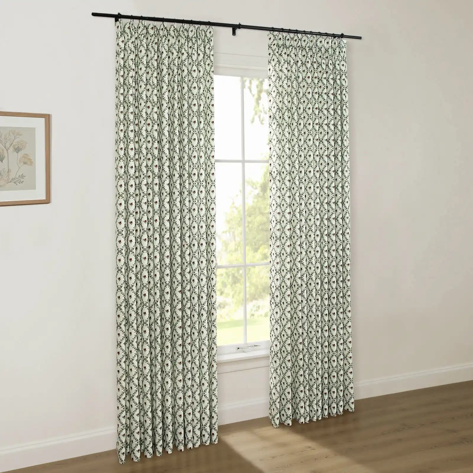 White pinch-pleat drapes with a dark green ogee pattern and red floral accents hang from a black rod, framing a window with lush green trees visible outside, in a room with a light wood floor and a framed botanical print
