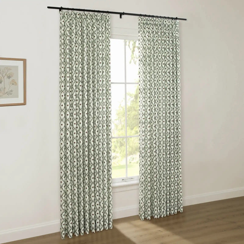 White pinch-pleat drapes with a dark green ogee pattern and red floral accents hang from a black rod, framing a window with lush green trees visible outside, in a room with a light wood floor and a framed botanical print