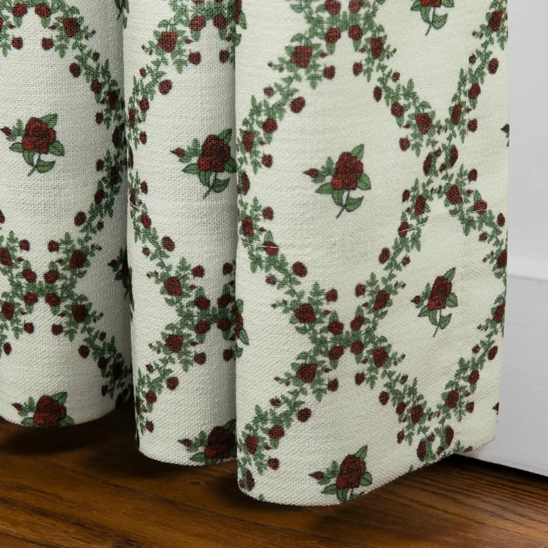 Textile print of a pleated off-white textured fabric curtain featuring a repeating pattern of deep red roses and green leafy vines forming diamond shapes above a dark wooden floor